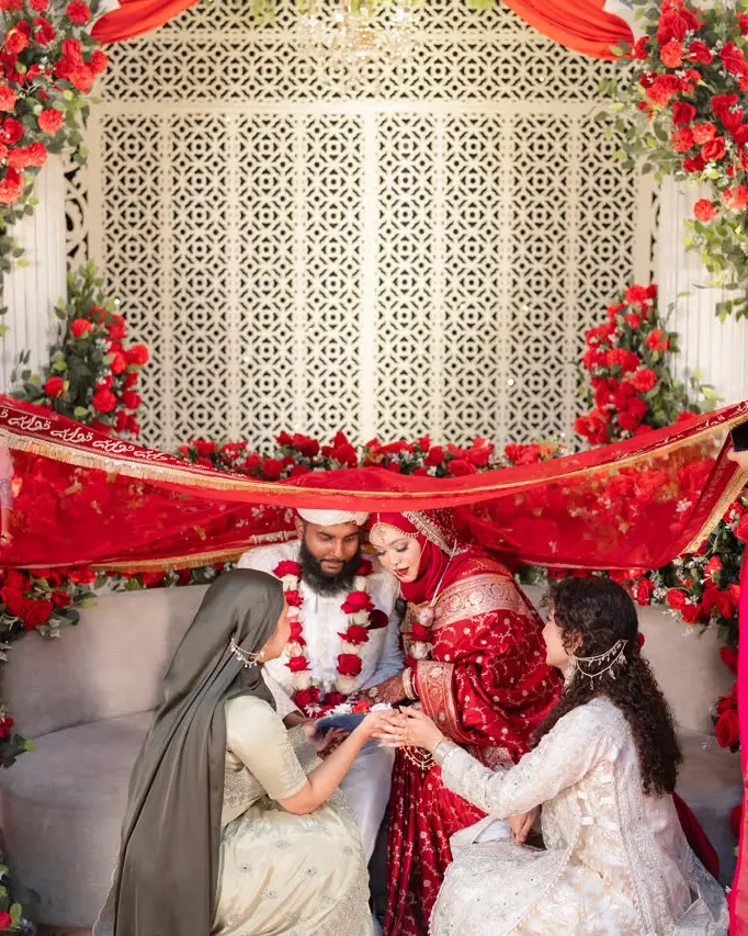 Family members gather around the couple on a floral stage, holding a red cloth above them