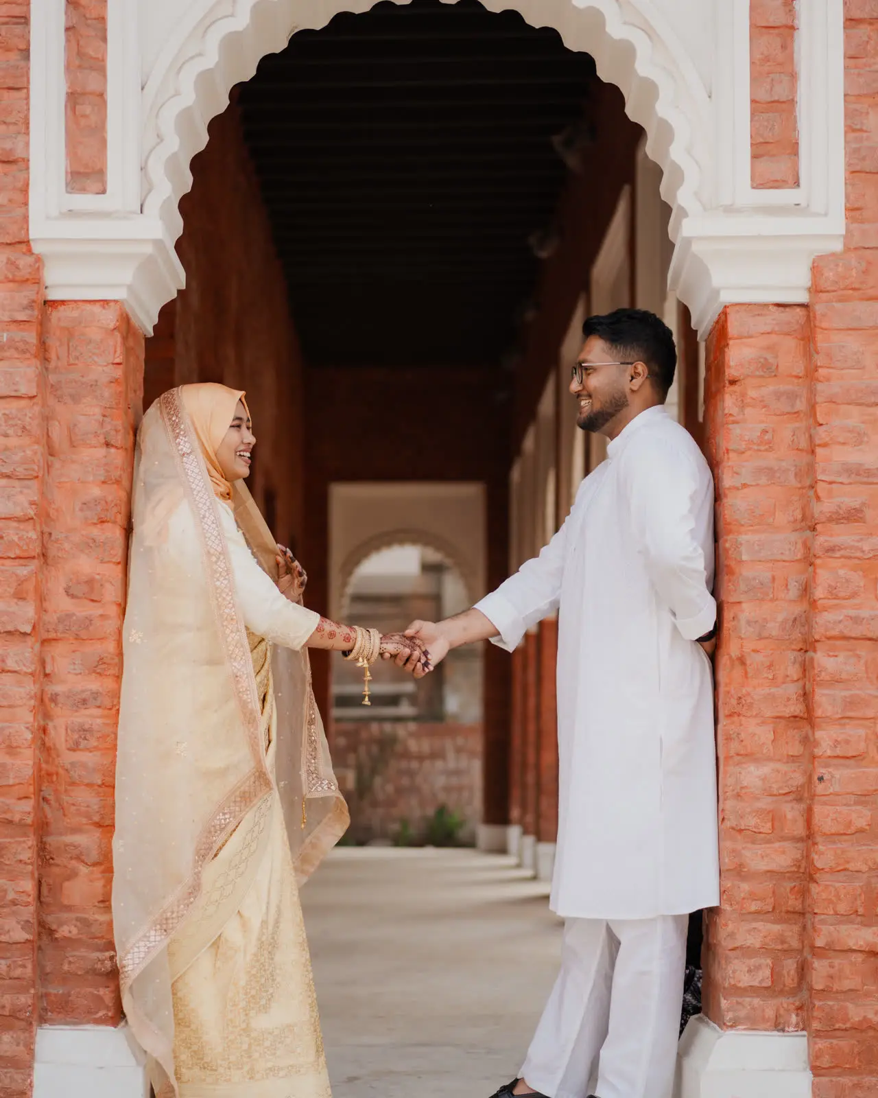 Anika and Faisal holding hands under a white arch in a brick corridor
