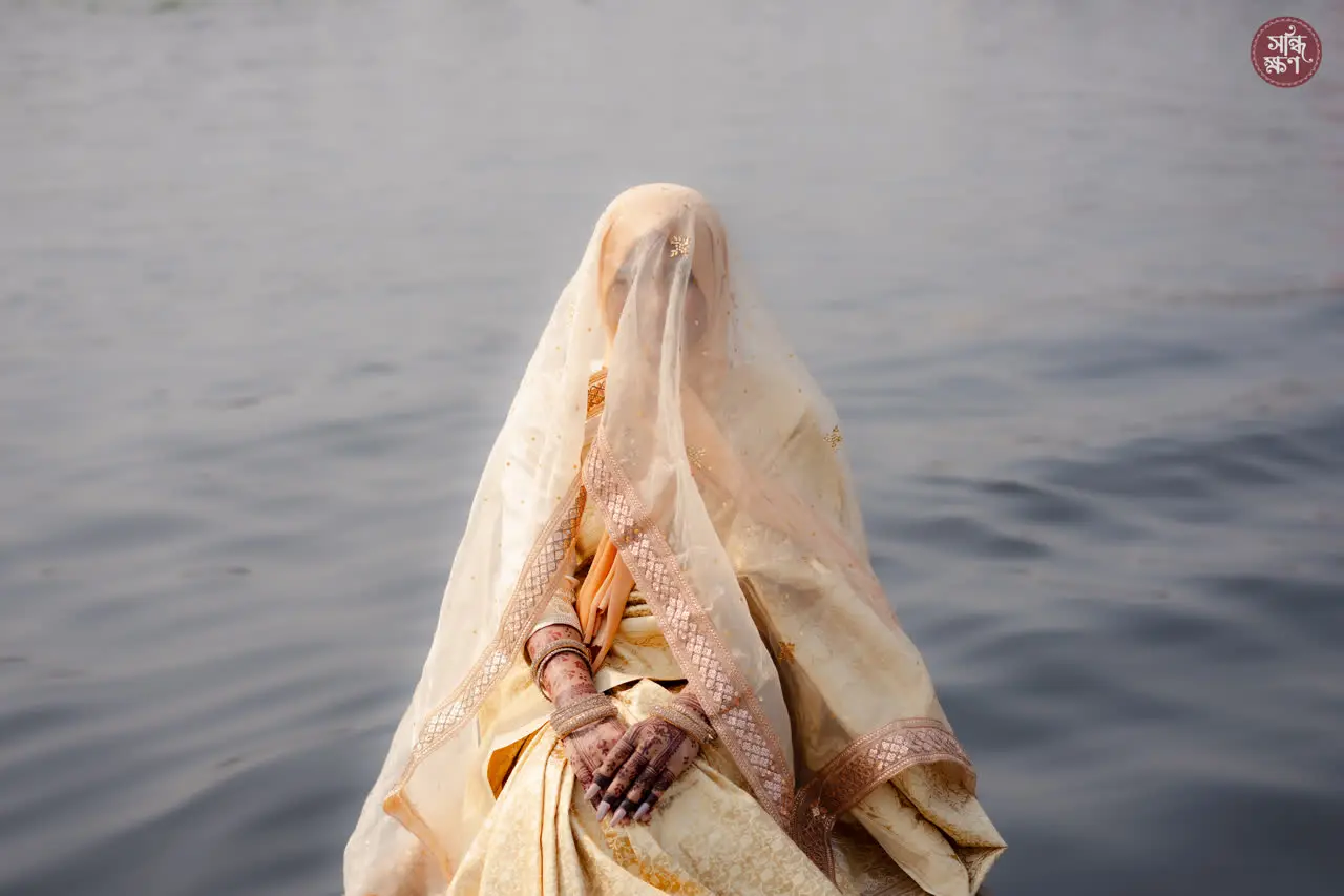 Nadia, veiled, sits at the edge of the water