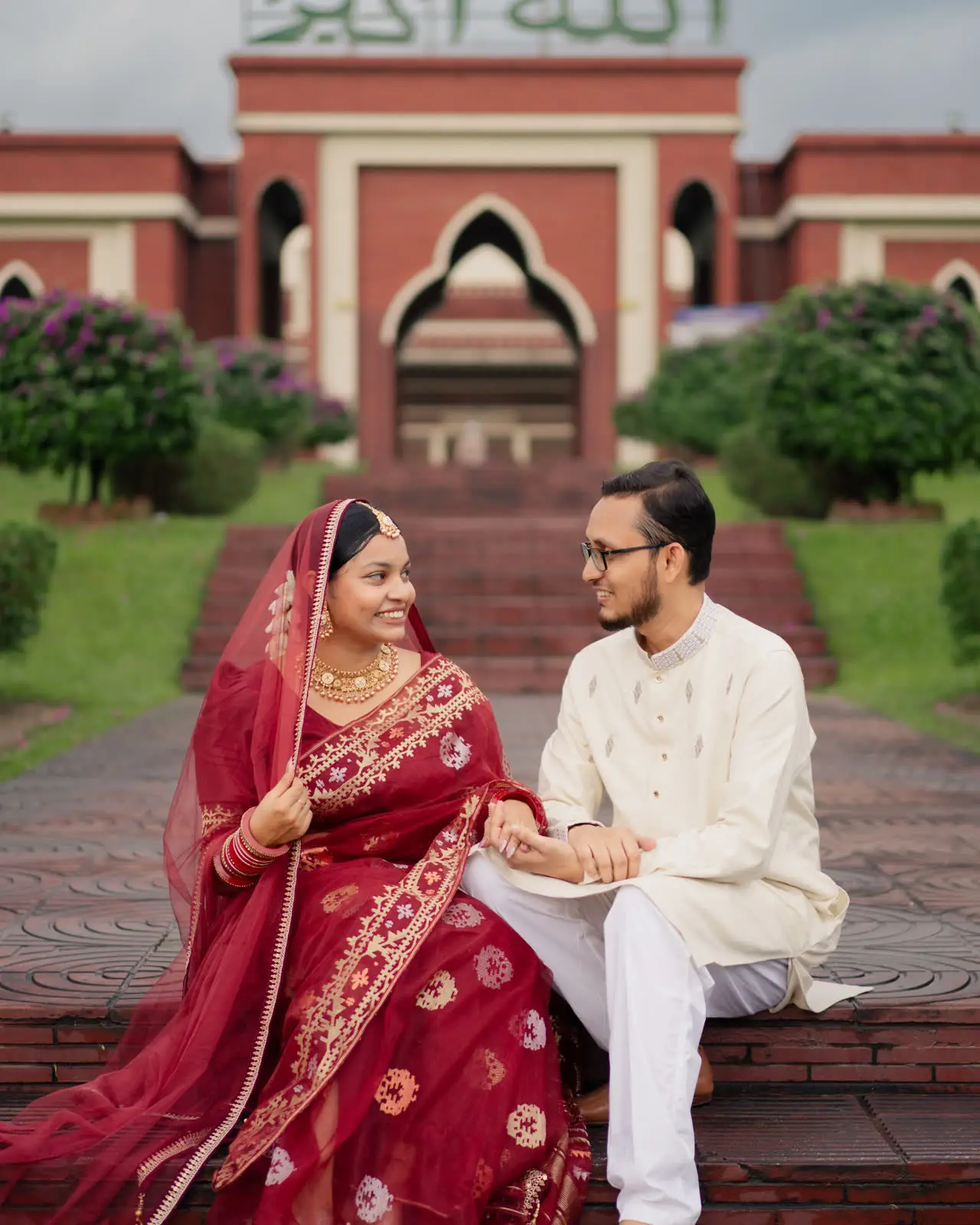 Sarah and Rahim sit on mosque steps, her red saree spread across the stone
