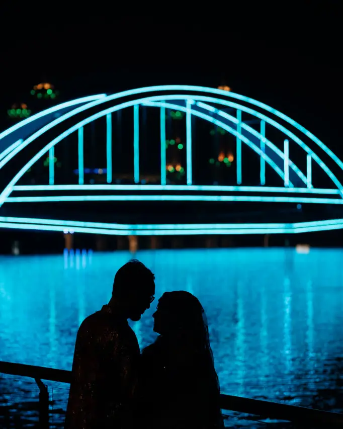 Rahim and Sarah in silhouette against a lit bridge reflected in the lake