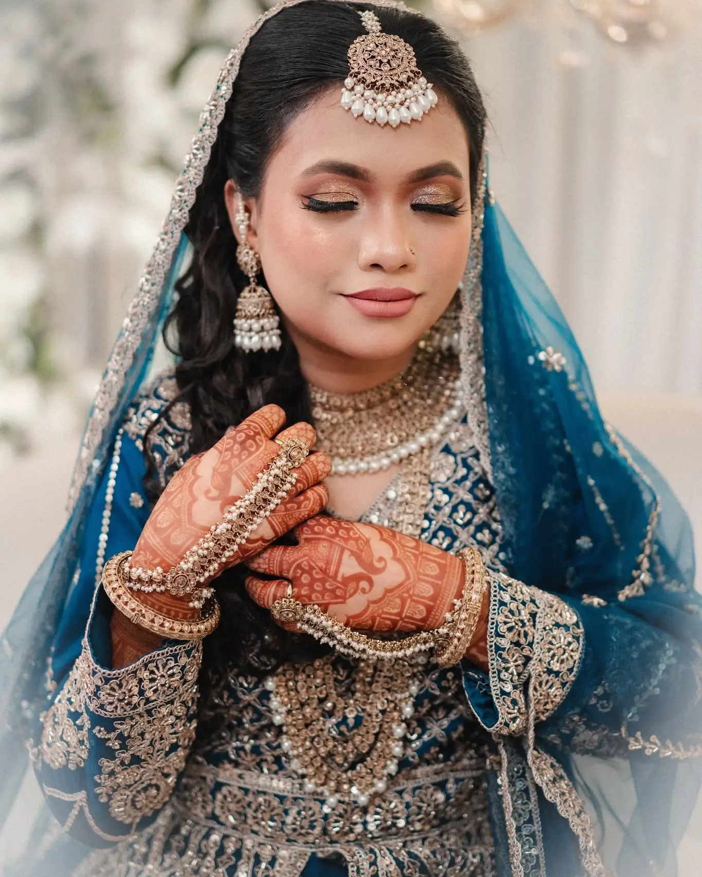 Portrait of the bride in a teal embroidered outfit and dupatta, with maang tikka, necklaces and hennaed hands.