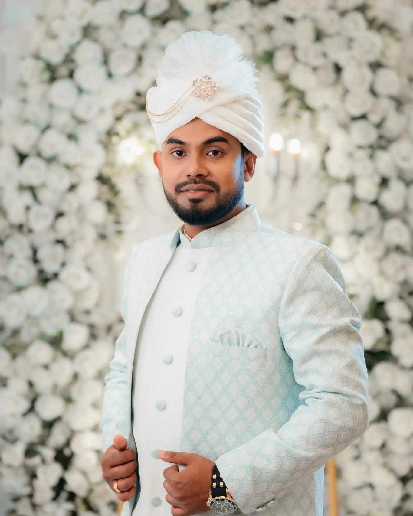 Portrait of the groom in a pale blue sherwani and white turban in front of a wall of white flowers.