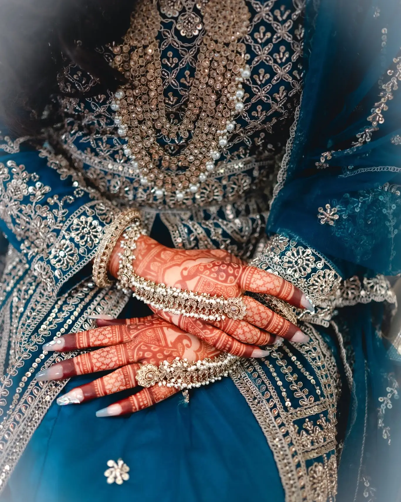Close-up of the bride's hennaed hands and rings resting on her teal outfit with silver embroidery.