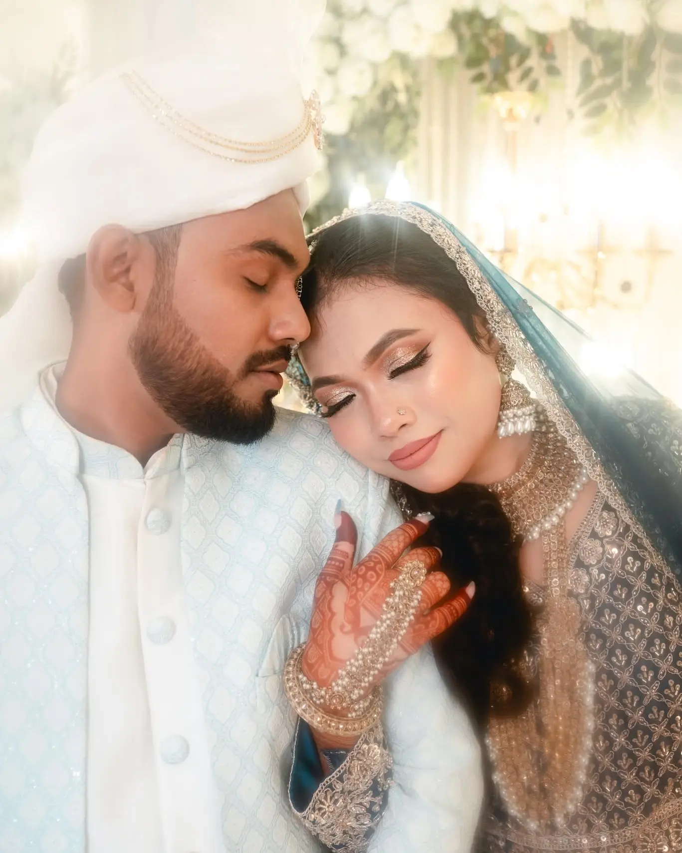 The couple cheek to cheek with eyes closed, the bride's hennaed hand on the groom's chest, soft lights behind.