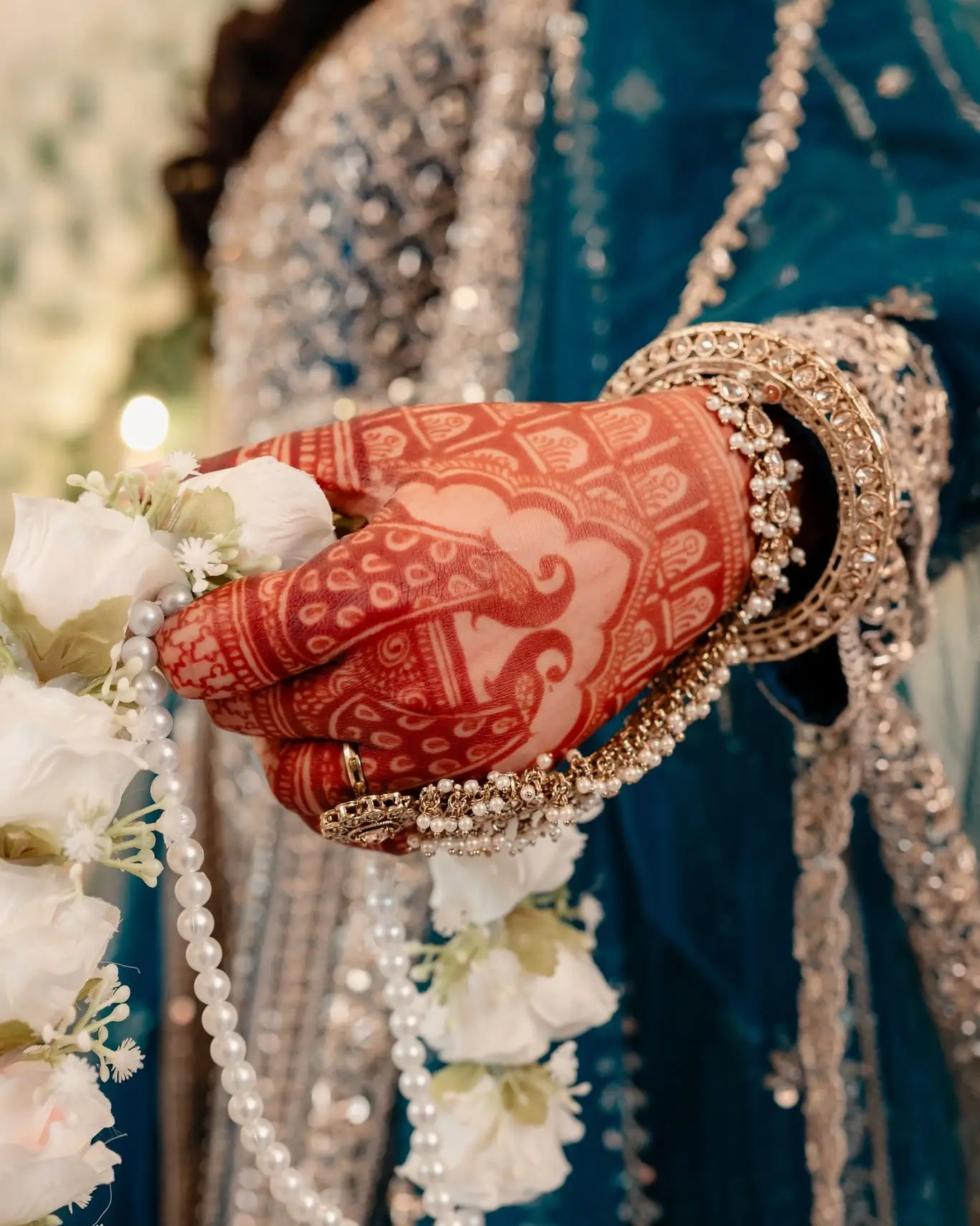 Close-up of the bride's hennaed hand and bangles holding a white flower garland with strands of pearls.