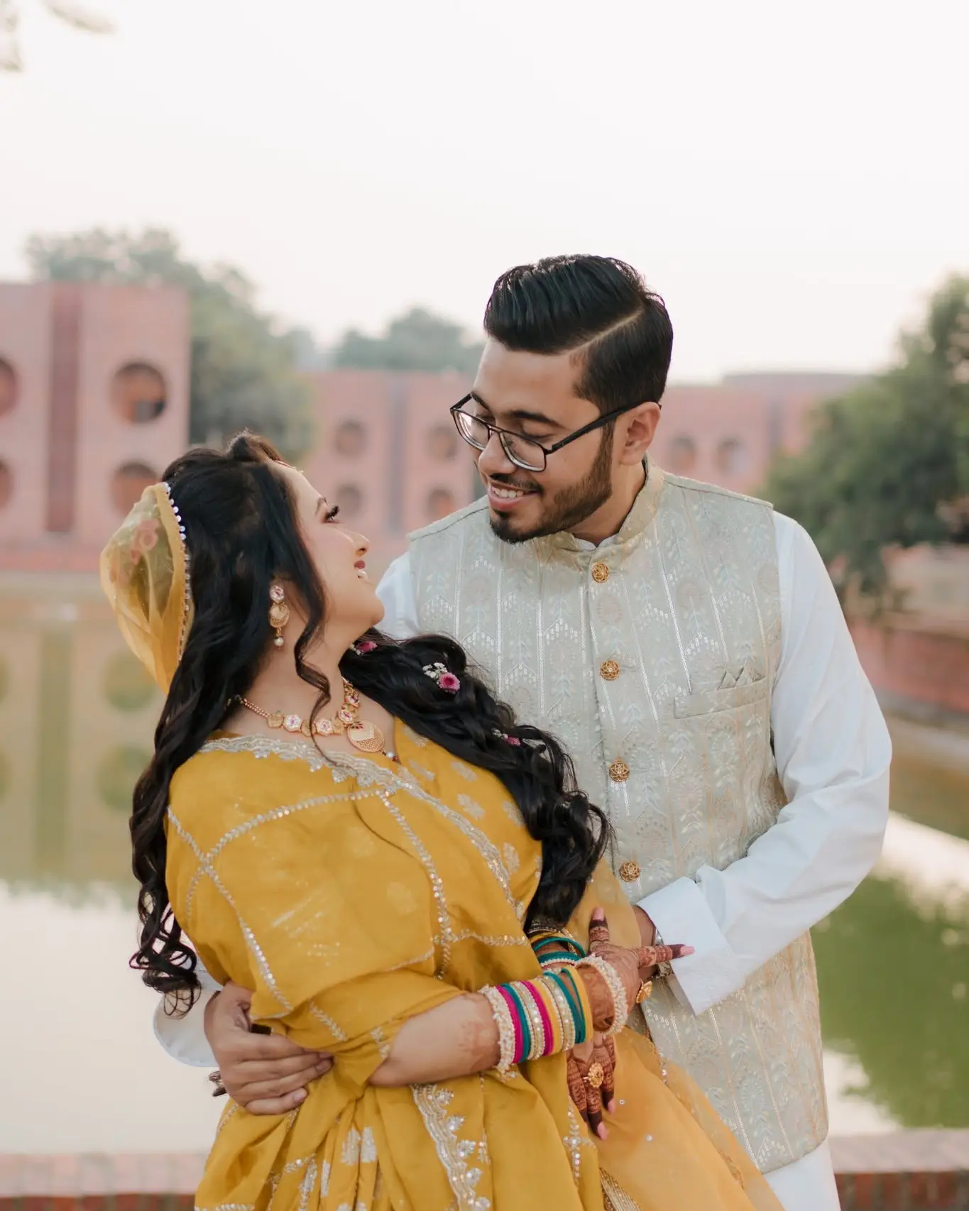 The bride in a mustard-yellow lehenga leans back laughing in the groom's arms beside a lake, red-brick buildings behind.