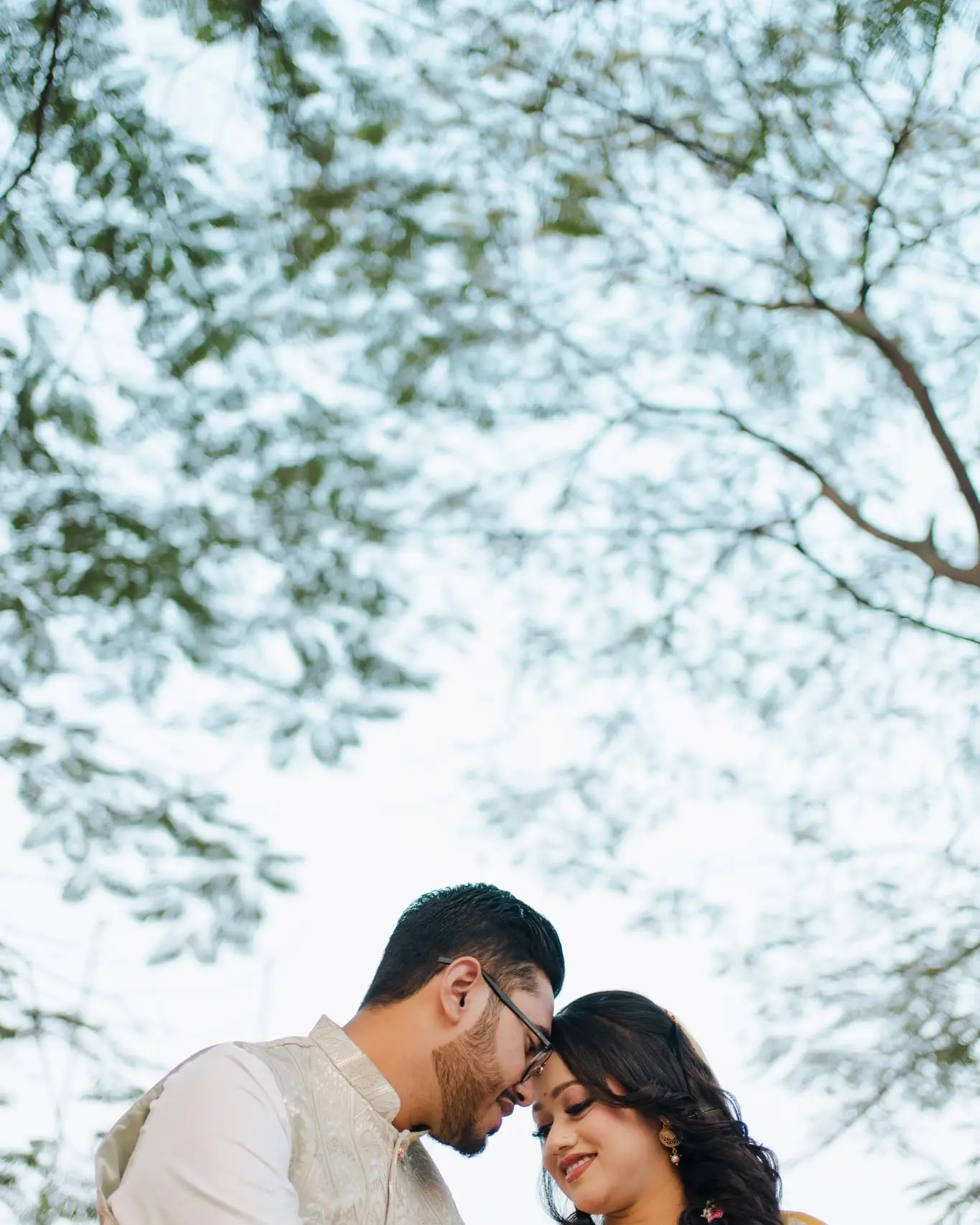 Low-angle view of the couple touching foreheads under leafy branches, the bride in yellow and the groom in a cream waistcoat.