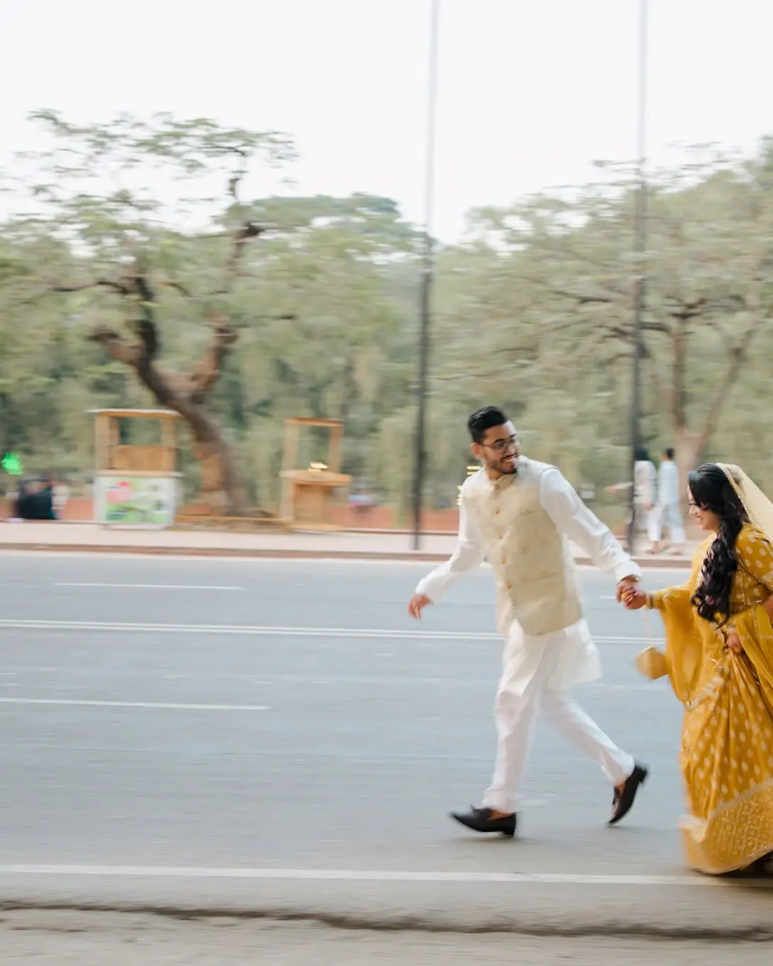 The couple run hand in hand across a wide, empty road lined with trees, the bride in yellow and the groom in white.
