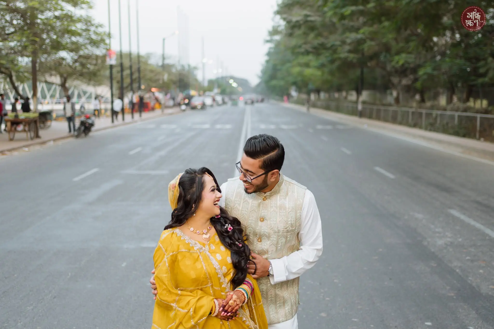 The couple laugh together on an empty tree-lined road, the groom embracing the bride from behind.