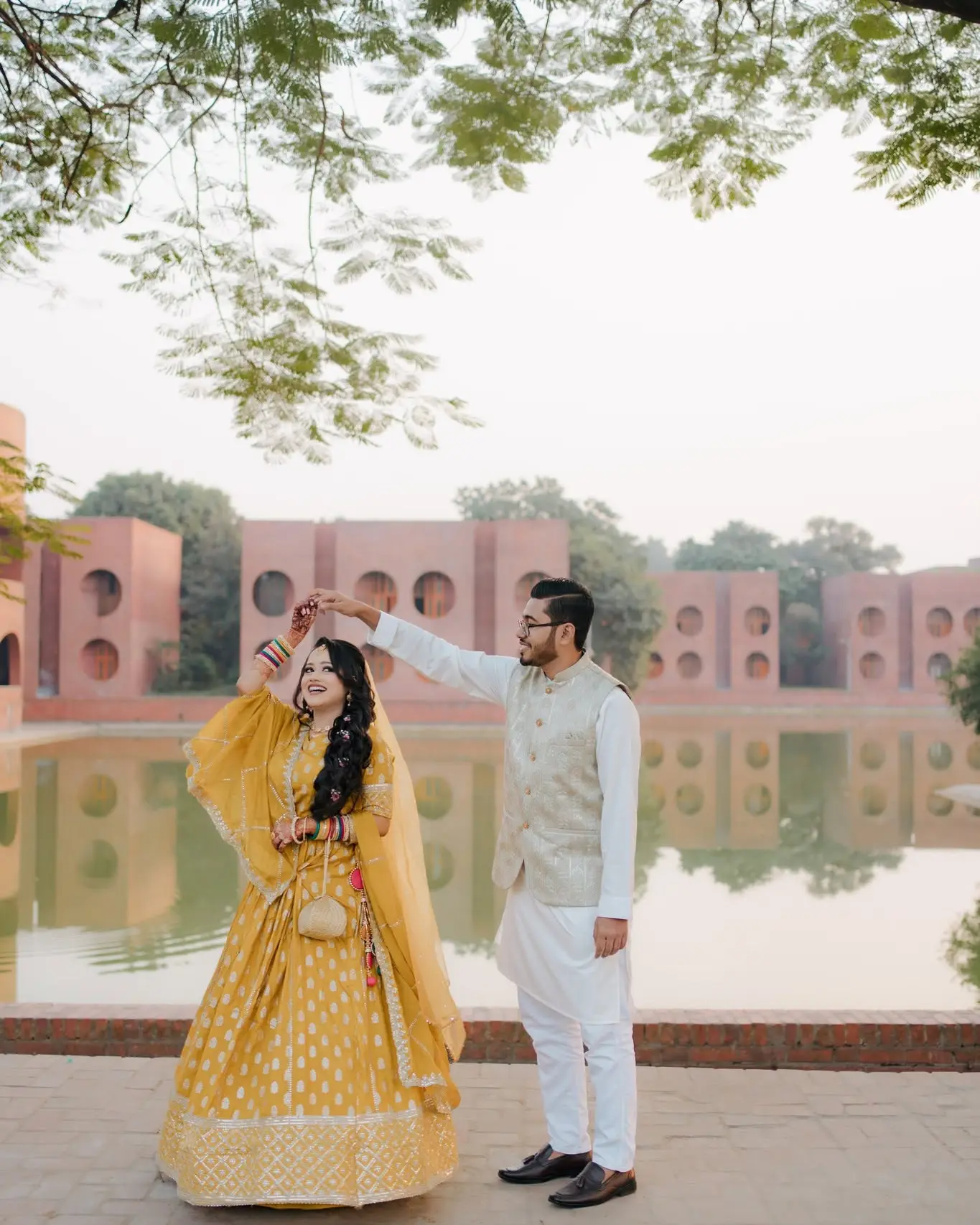 The groom twirls the bride in her yellow lehenga beside a lake, red-brick buildings with round openings behind.